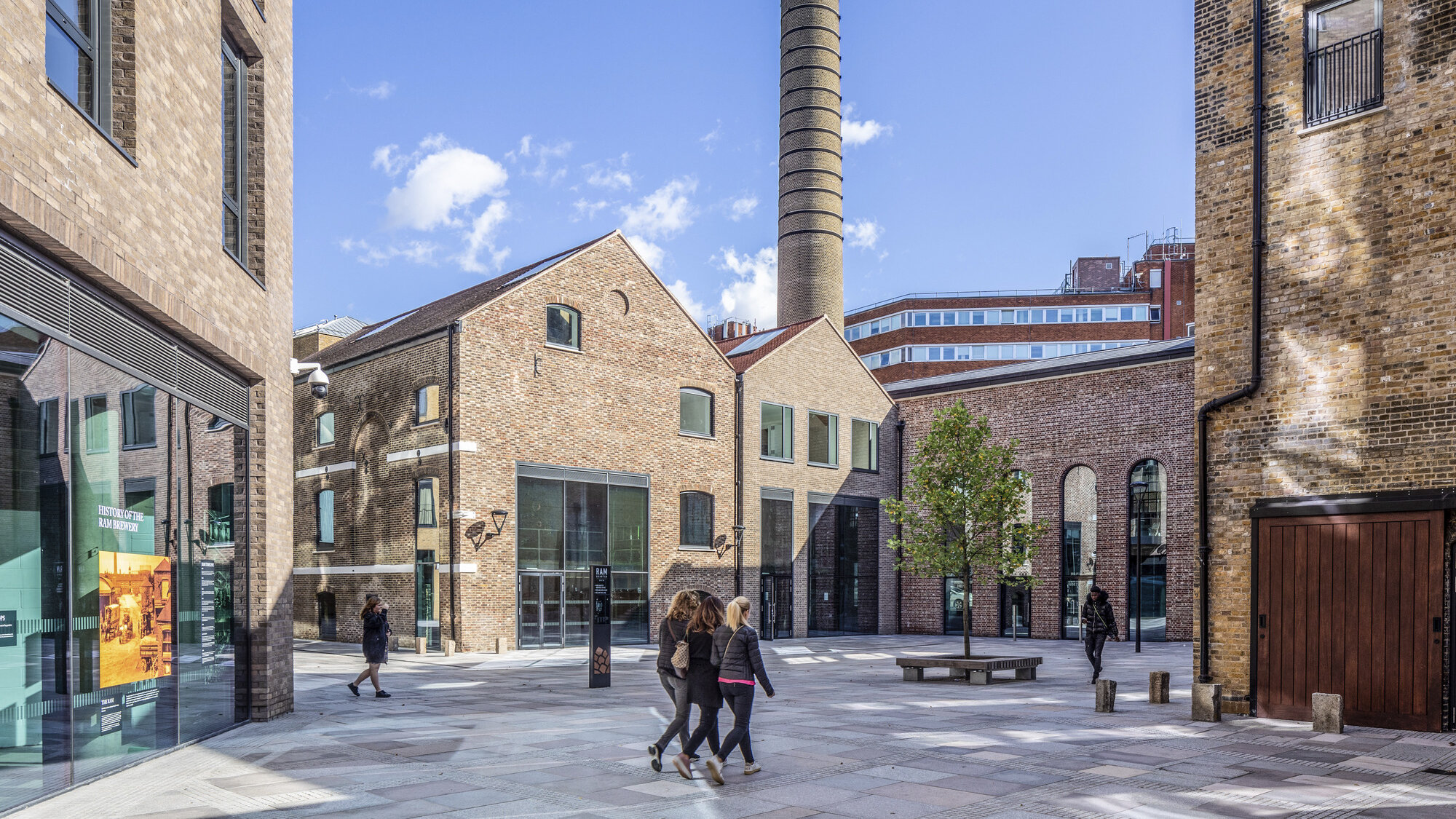 Public courtyard at Young's Ram Brewery with historic brick buildings, smokestack, urban trees, and pedestrian-friendly landscape architecture.