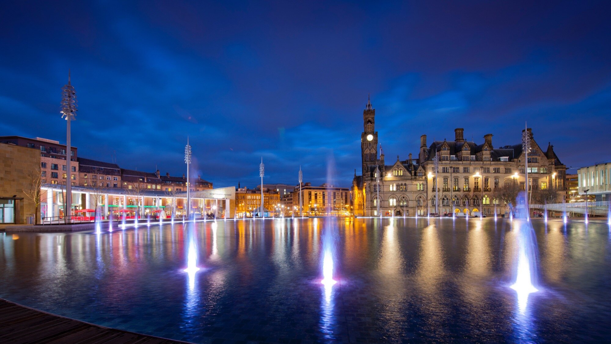 Illuminated city square with modern fountain water feature and historic architecture, showcasing urban landscape design and public realm lighting at Bradford City Park, designed by Yorkshire based studio, Gillespies.