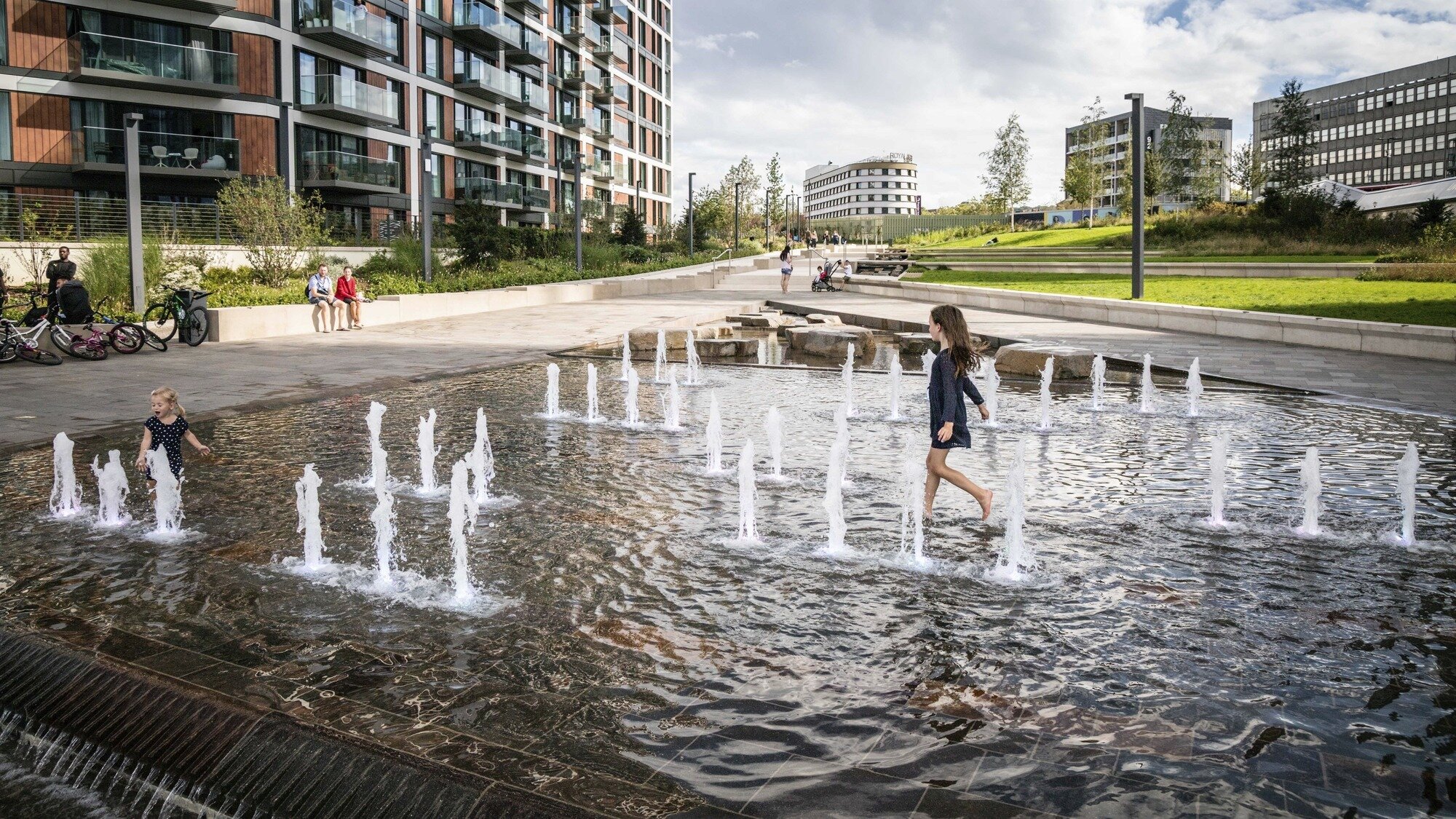 Children playing in interactive water fountain at Royal Arsenal Riverside waterfront urban landscape design.