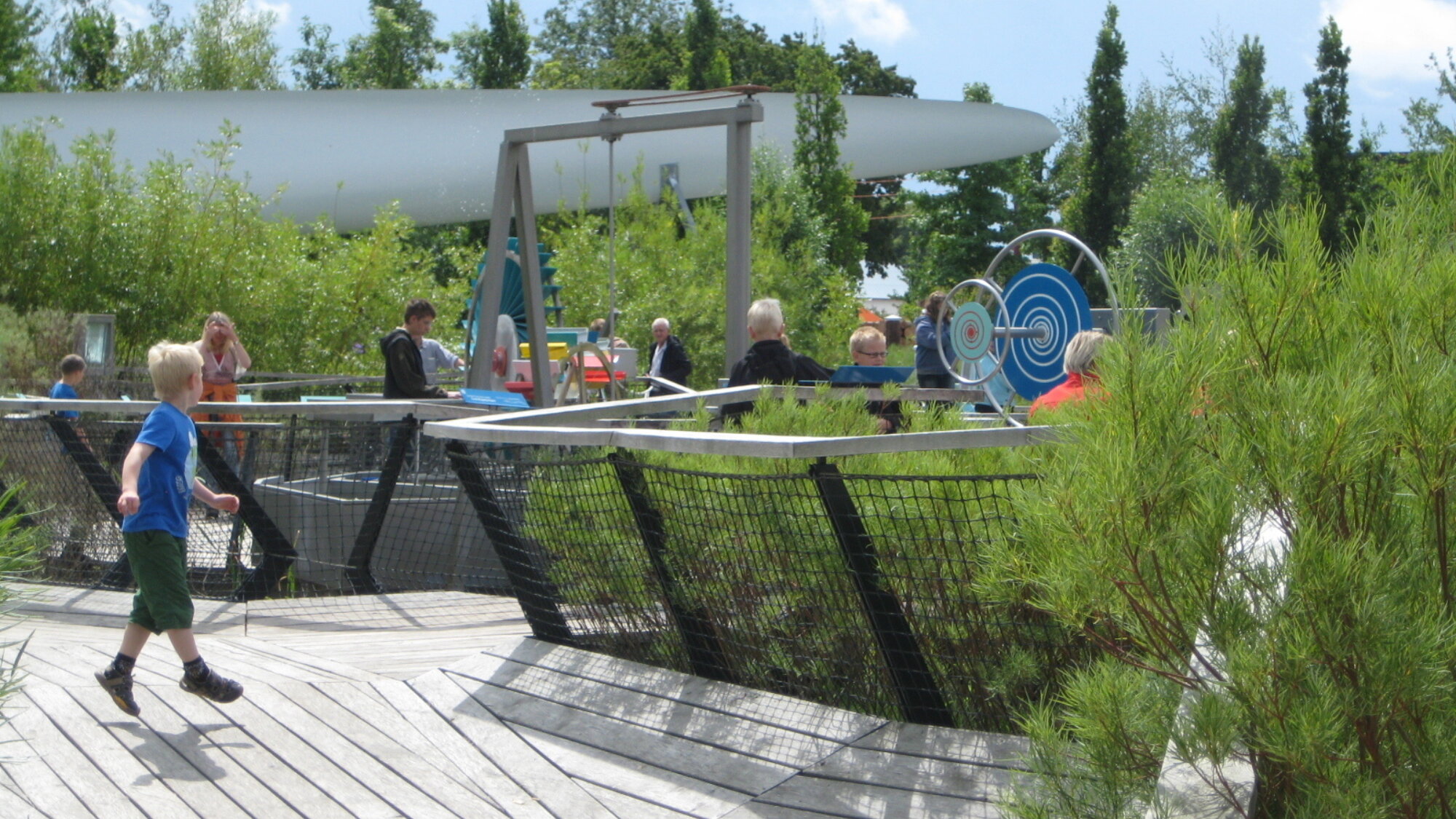 Children and adults explore interactive outdoor science exhibits at Universe Science Park, with timber decking, planted beds and hands-on mechanical installations set within a landscaped garden.