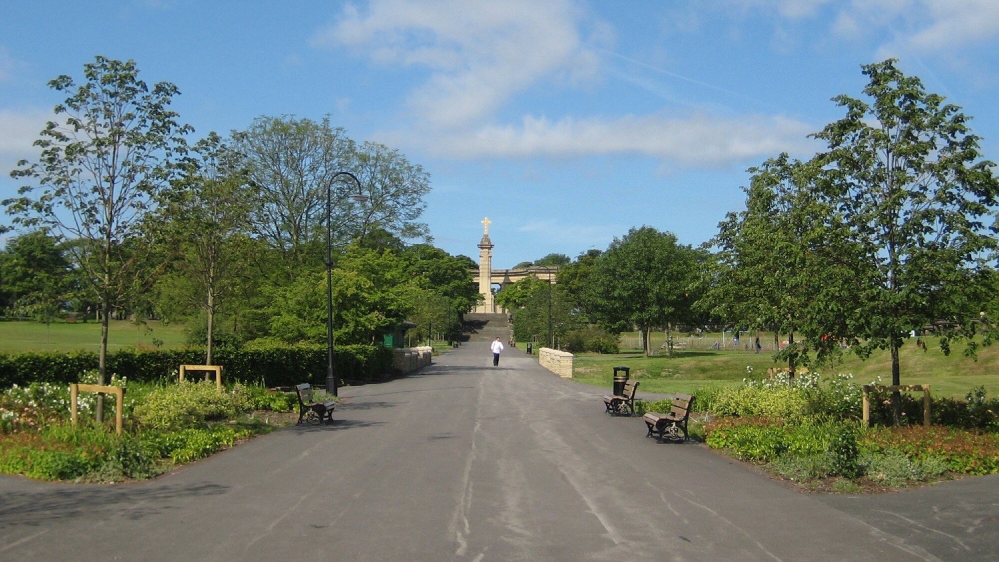 Tree-lined promenade in Greenhead Park with benches, flower beds, and landscaped pathways leading to historic monument.