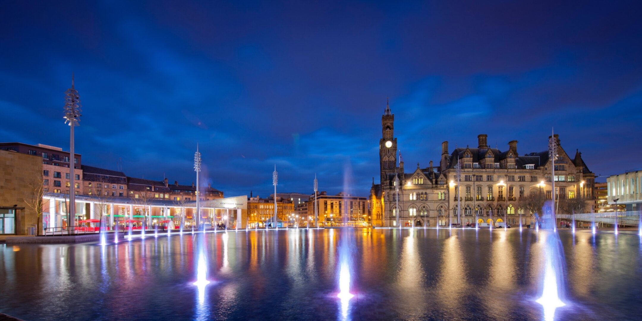 Illuminated city square with modern fountain water feature and historic architecture, showcasing urban landscape design and public realm lighting at Bradford City Park, designed by Yorkshire based studio, Gillespies.