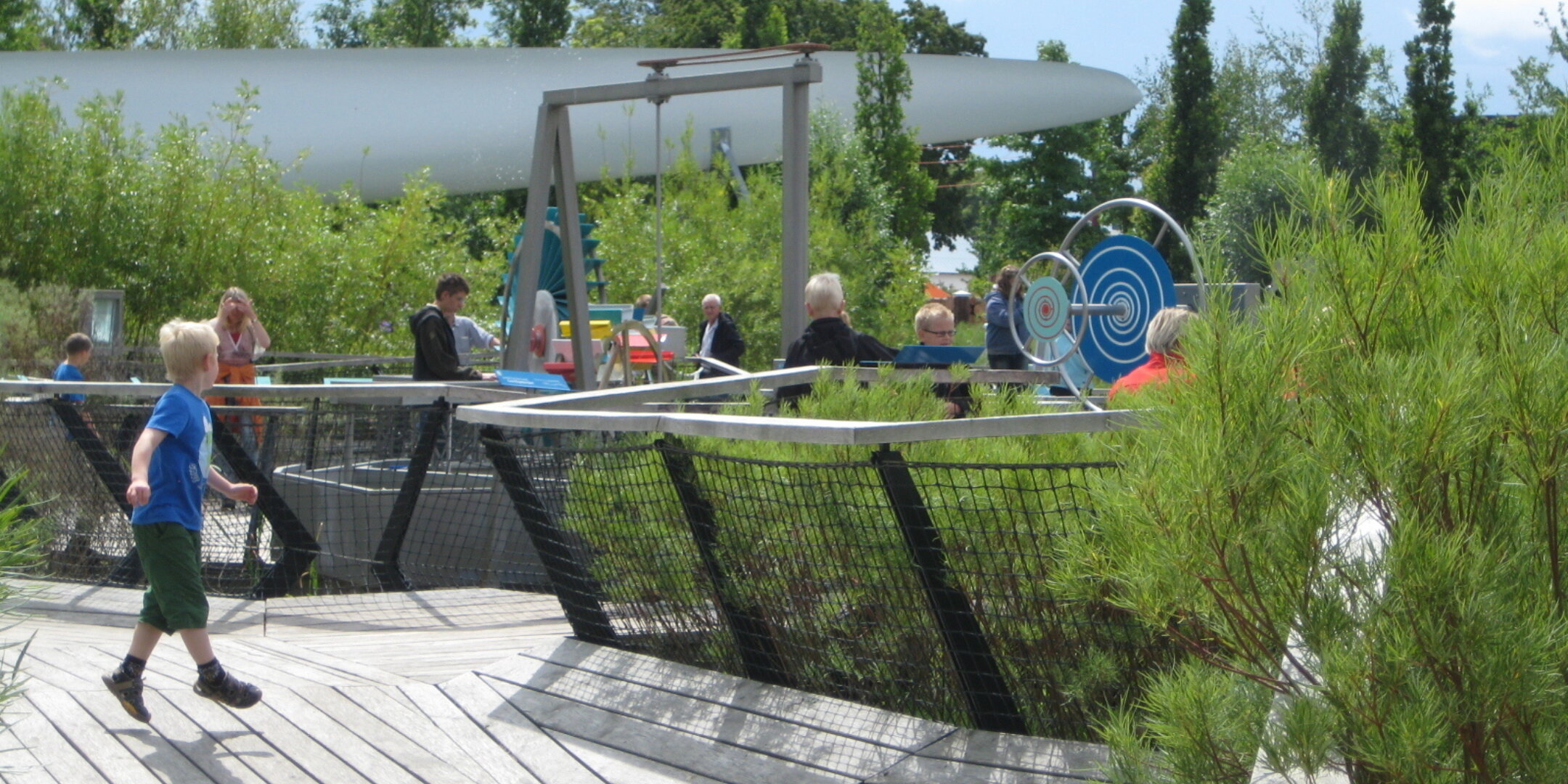 Children and adults explore interactive outdoor science exhibits at Universe Science Park, with timber decking, planted beds and hands-on mechanical installations set within a landscaped garden.