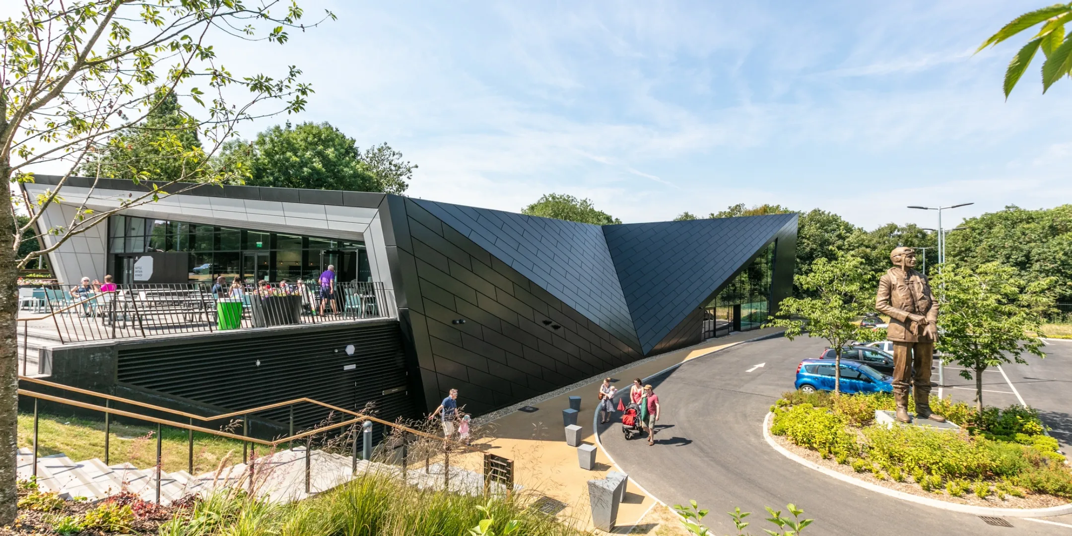 The folded geometry of the visitor centre emerges from a landscaped setting of woodland planting and sweeping paths, creating a strong architectural presence within a calm, green backdrop.