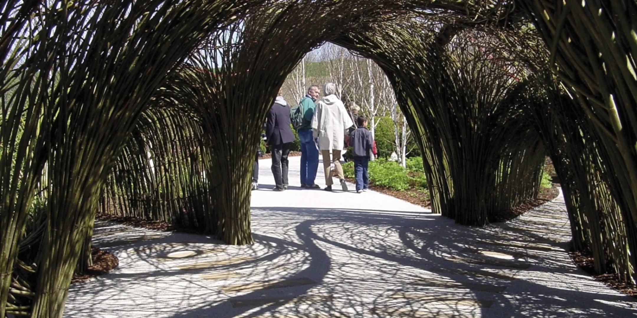 Arched woven willow tunnel at Universe Science Park Danfoss, casting intricate shadows across a paved pathway as visitors walk through landscaped gardens