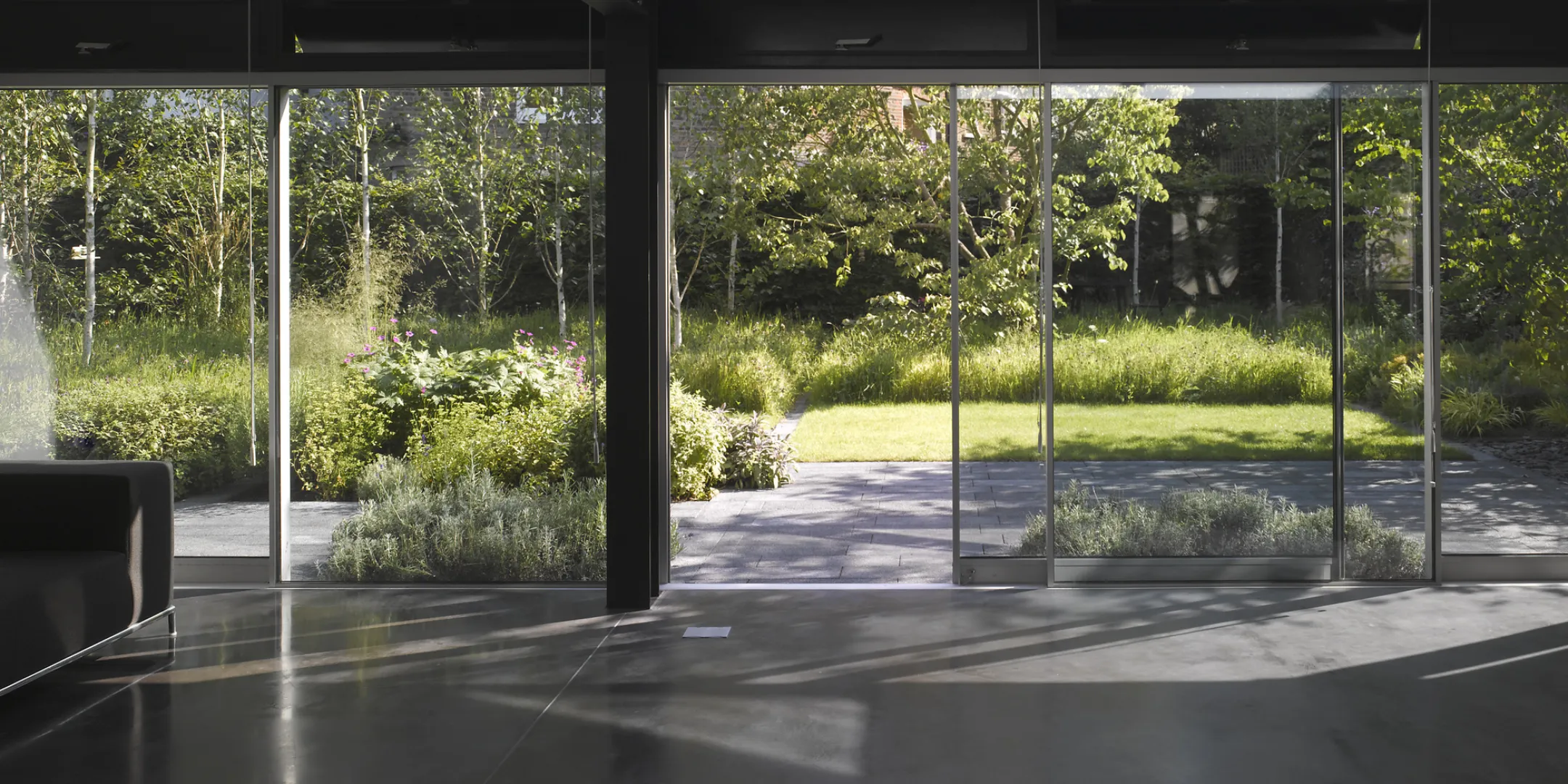Floor to ceiling sliding glass doors open onto a landscaped garden with lawn, trees, and layered planting beds, viewed from a modern interior with dark structural beams and a paved terrace in the foreground.