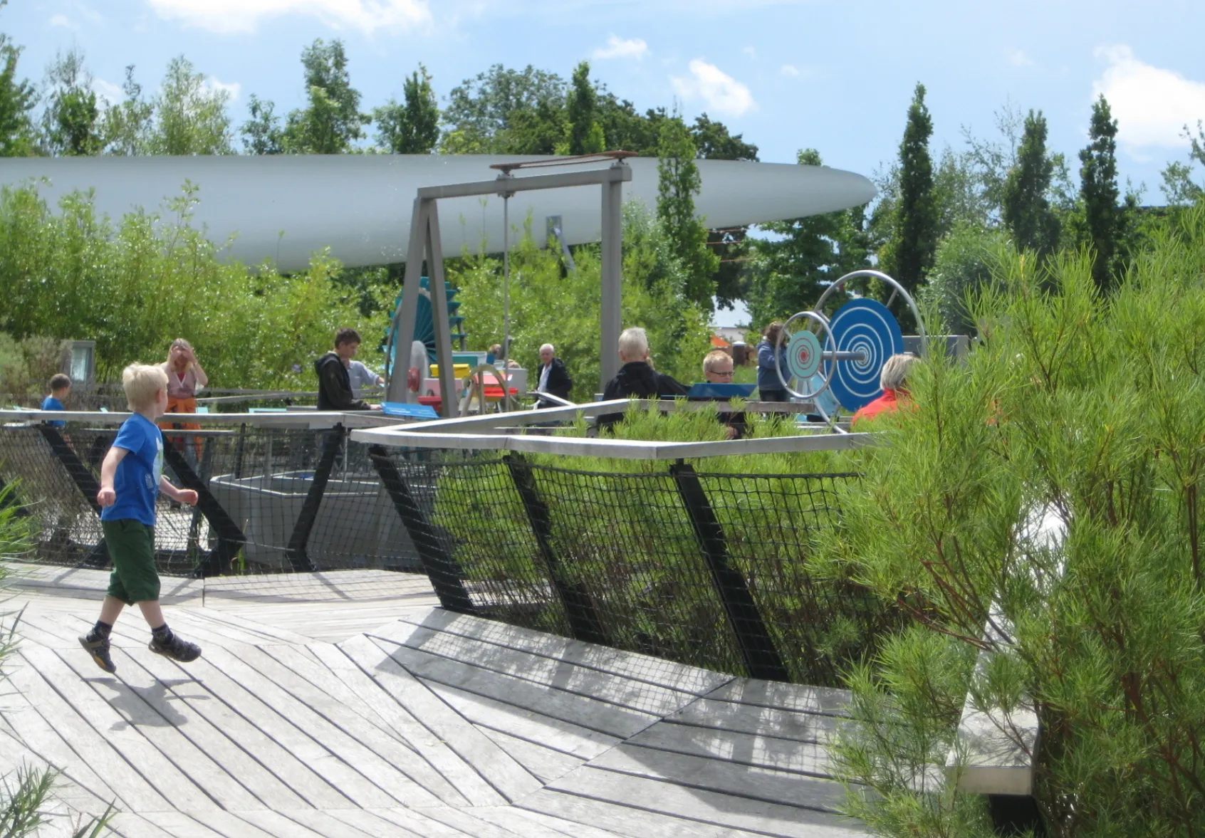 Children and adults explore interactive outdoor science exhibits at Universe Science Park, with timber decking, planted beds and hands-on mechanical installations set within a landscaped garden.