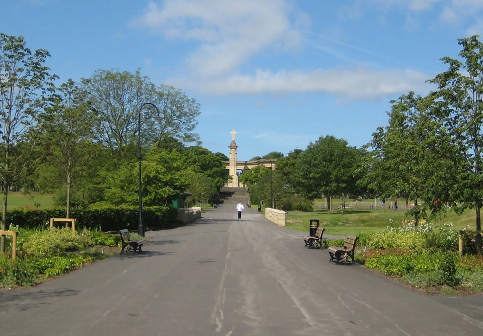 Tree-lined promenade in Greenhead Park with benches, flower beds, and landscaped pathways leading to historic monument.