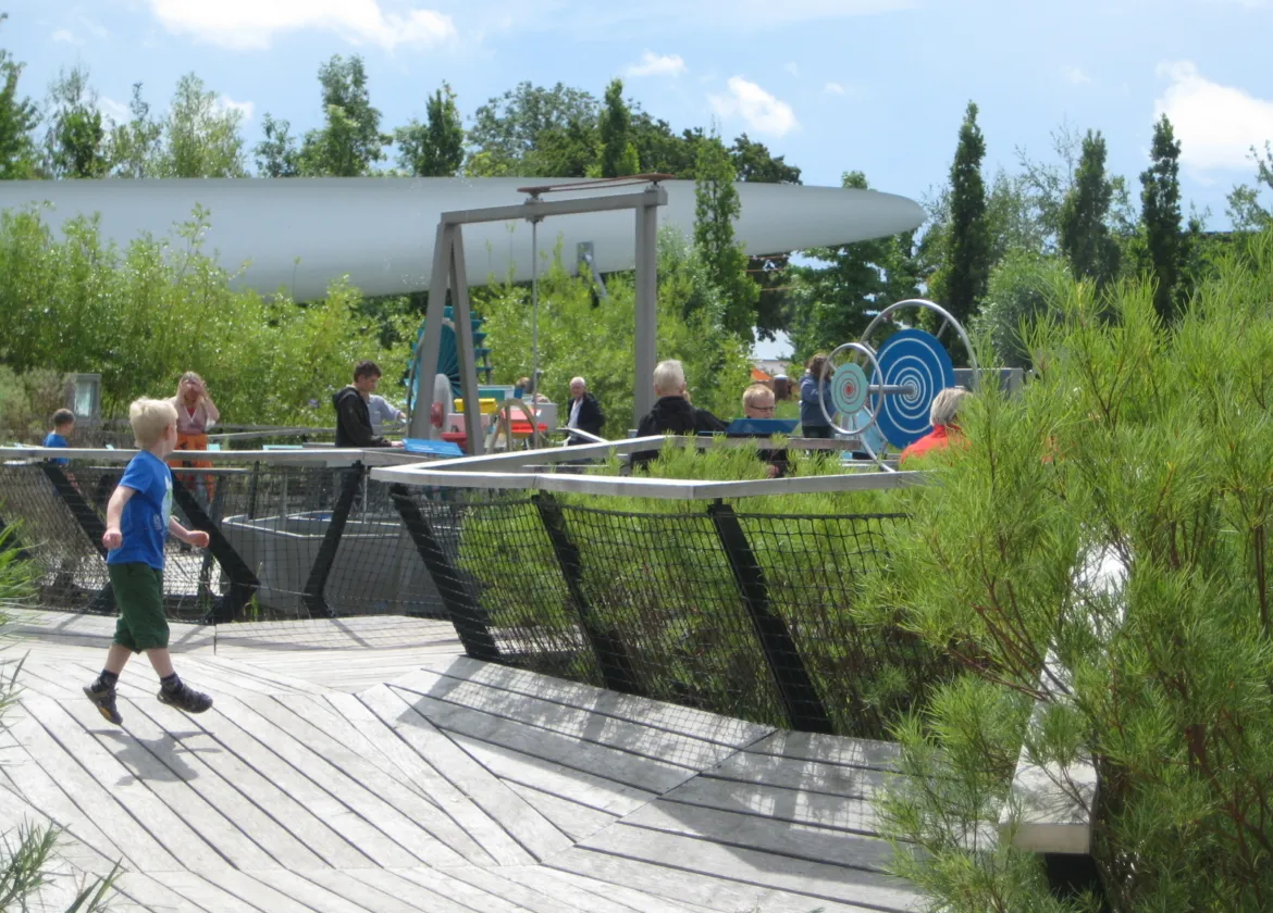 Children and adults explore interactive outdoor science exhibits at Universe Science Park, with timber decking, planted beds and hands-on mechanical installations set within a landscaped garden.