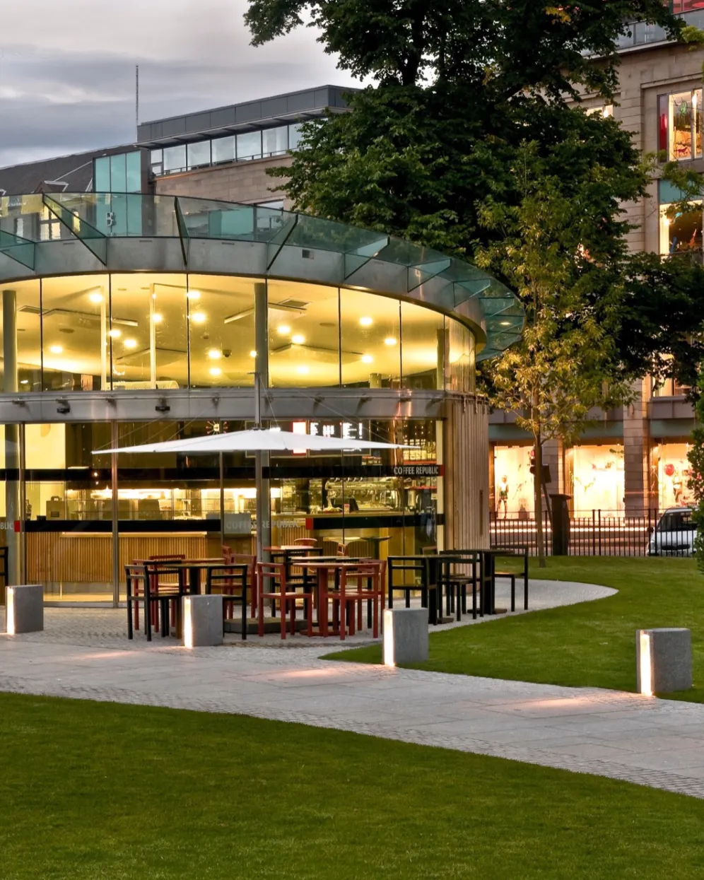 Modern glass café pavilion in St Andrew Square surrounded by lawn, lighting, and contemporary urban landscape design. Landscape and pavilion architecture designed by renowned design firm, Gillespies.