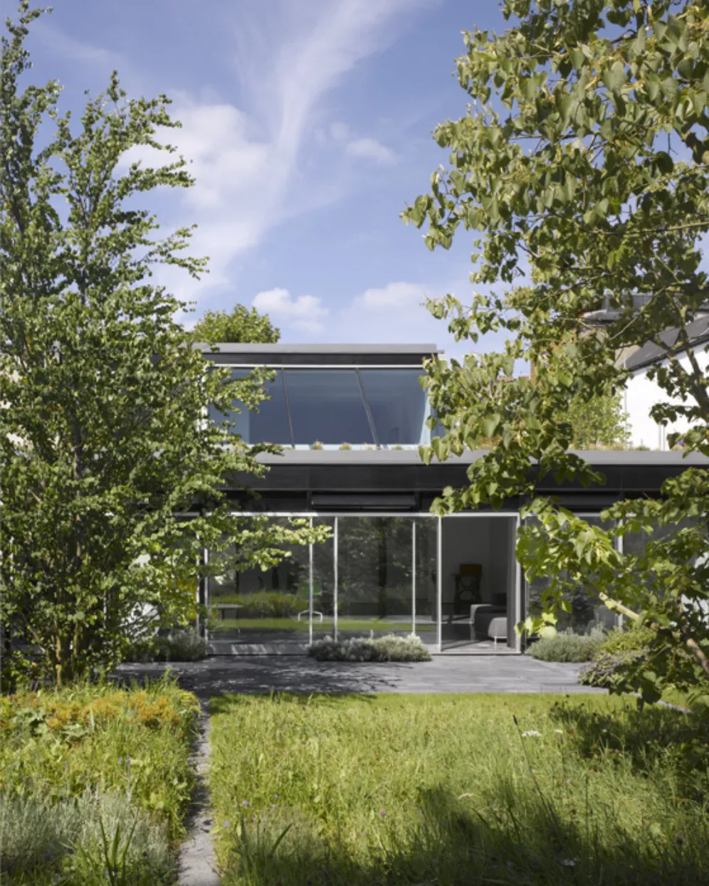Modern two storey house with expansive sliding glass doors opening onto a lawn and planted garden, framed by mature trees and a paved terrace under a bright blue sky.