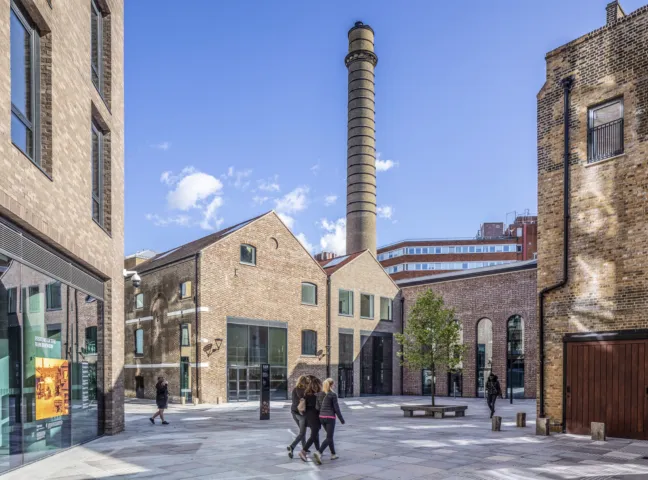 Public courtyard at Young's Ram Brewery with historic brick buildings, smokestack, urban trees, and pedestrian-friendly landscape architecture.