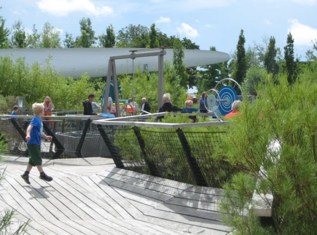 Children and adults explore interactive outdoor science exhibits at Universe Science Park, with timber decking, planted beds and hands-on mechanical installations set within a landscaped garden.