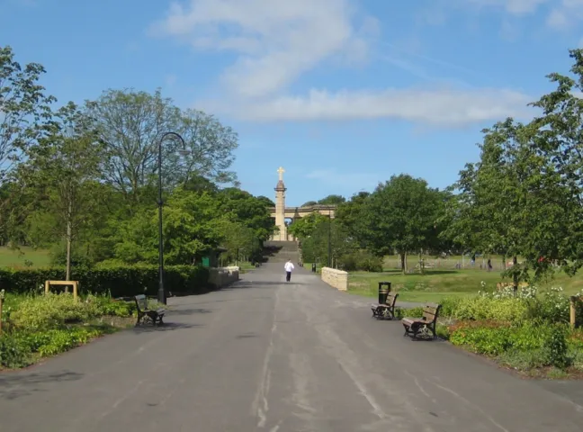 Tree-lined promenade in Greenhead Park with benches, flower beds, and landscaped pathways leading to historic monument.
