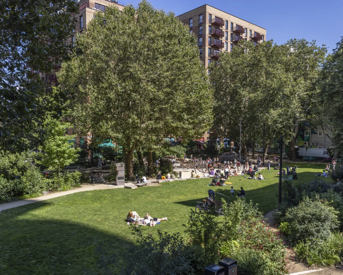 Residents and visitors gather on a sunlit lawn at Elephant Park, surrounded by mature trees, planting and new residential buildings.