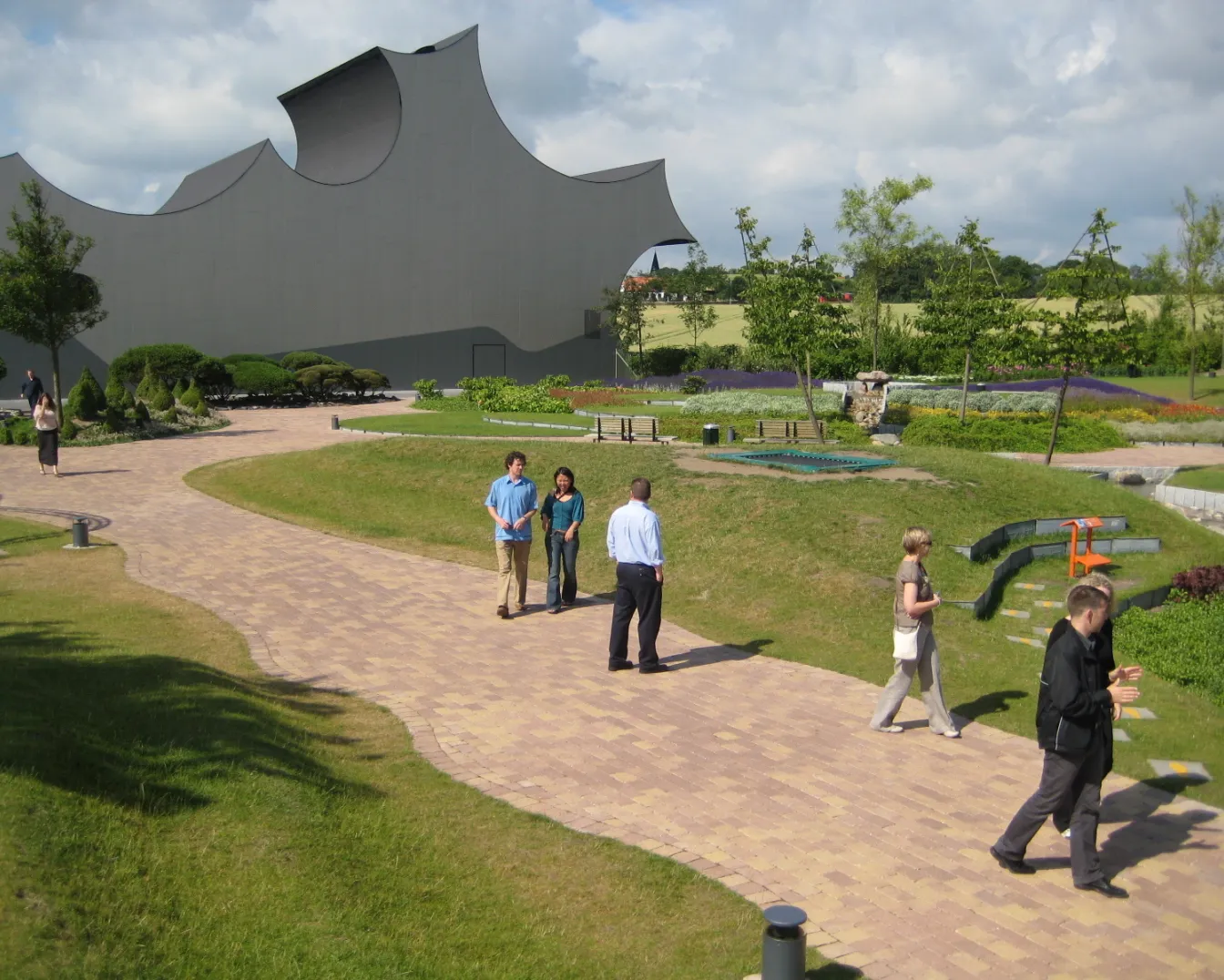 Curving paved pathways wind through landscaped gardens at Universe Science Park Danfoss, with visitors walking among lawns, planting beds and interactive outdoor exhibits beside a distinctive sculptural building
